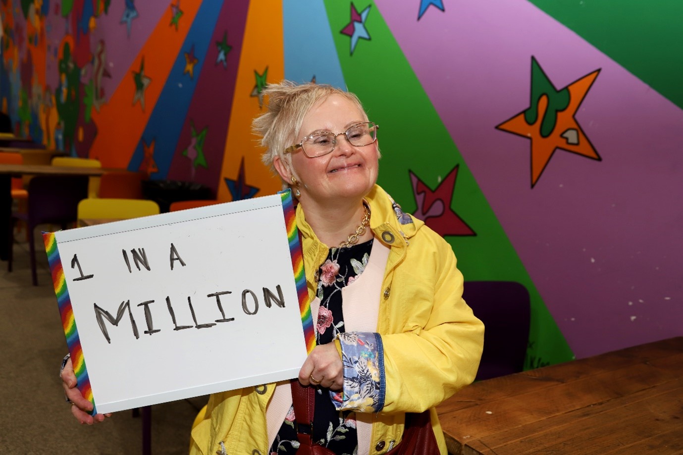 A photo of a woman in a yellow coat. She is wearing glasses and smiling. She is holding up a whiteboard with the words '1 in a Million' written on it in black ink.