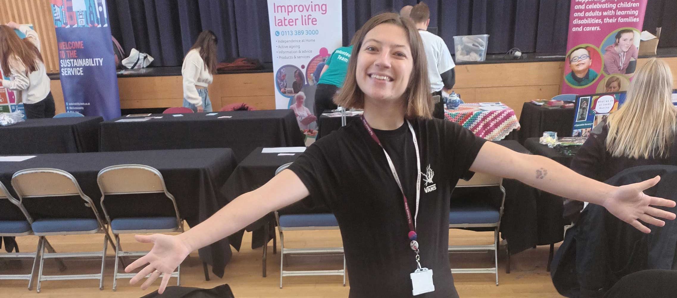 A photo of Beth, Leeds Autism AIM's Volunteer Coordinator, at a stall for a volunteering event. They have their arms outstretched. They are also smiling.