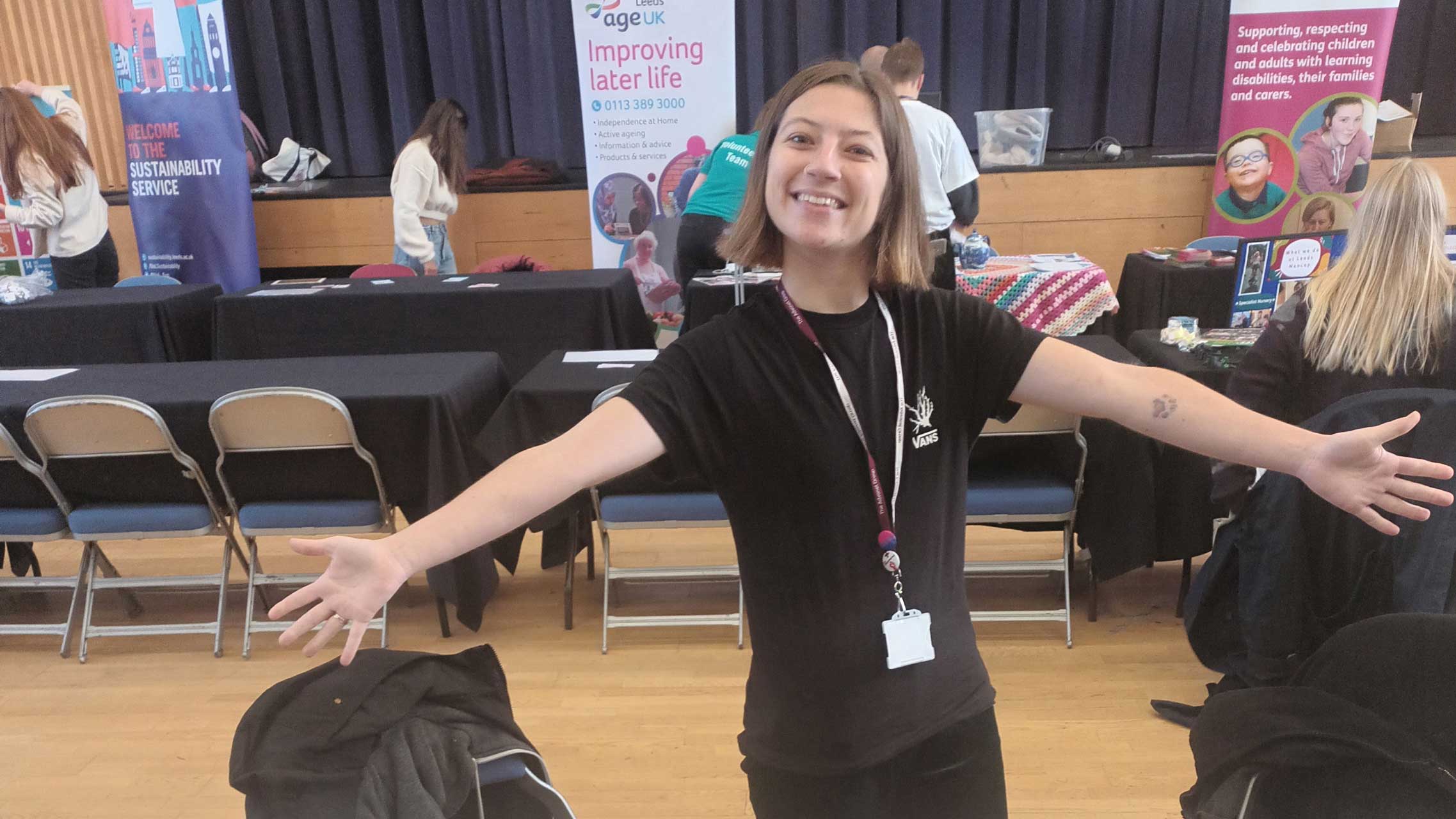 A photo of Beth, Leeds Autism AIM's Volunteer Coordinator, at a stall for a volunteering event. They have their arms outstretched. They are also smiling.