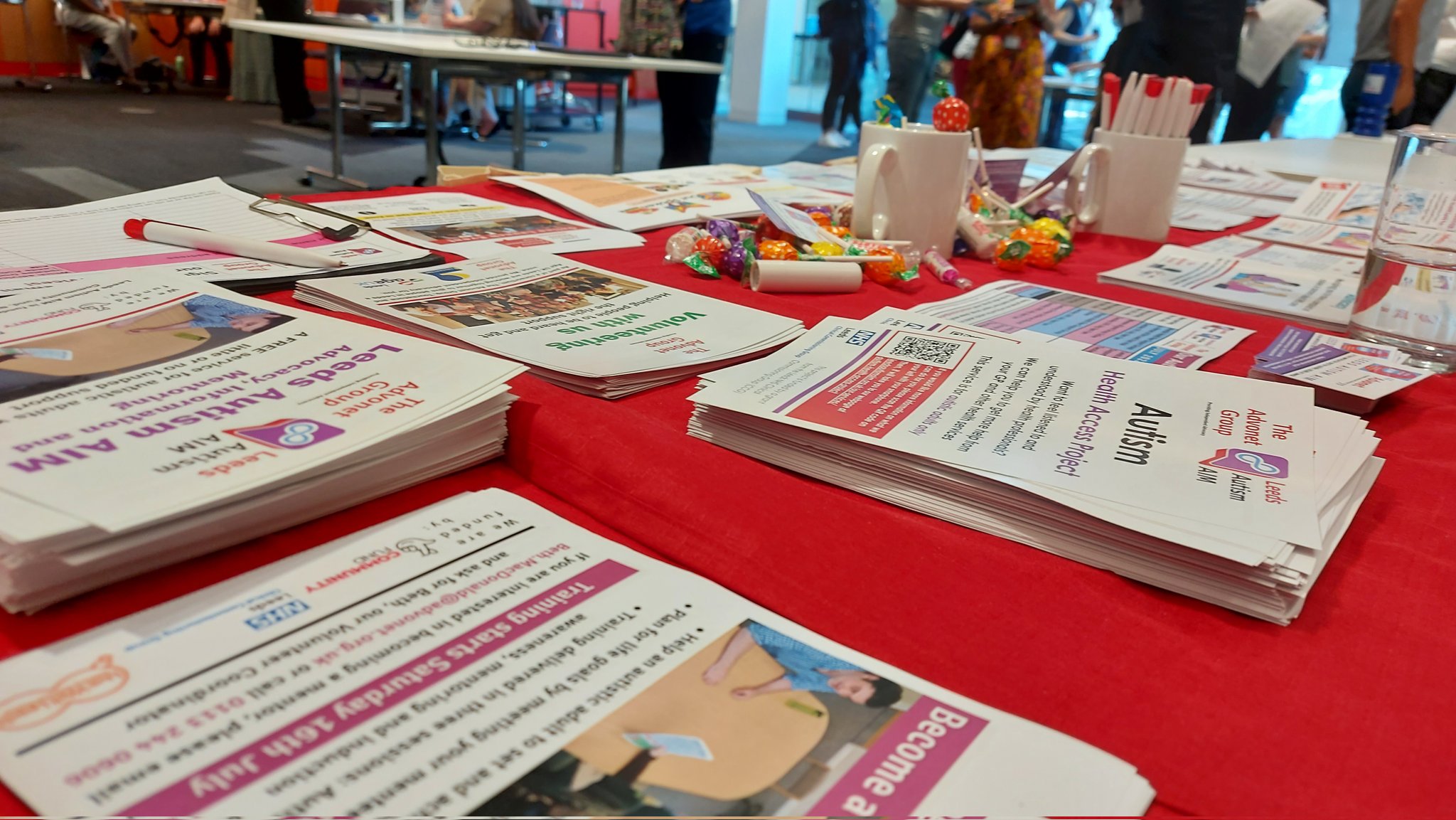 A photo of a table with a red tablecloth on it. On top of the tablecloth, there are leaflets and flyers for our Leeds Autism AIM service, as well as other leaflets and business cards, plus a couple of mugs with sweets and pens inside.