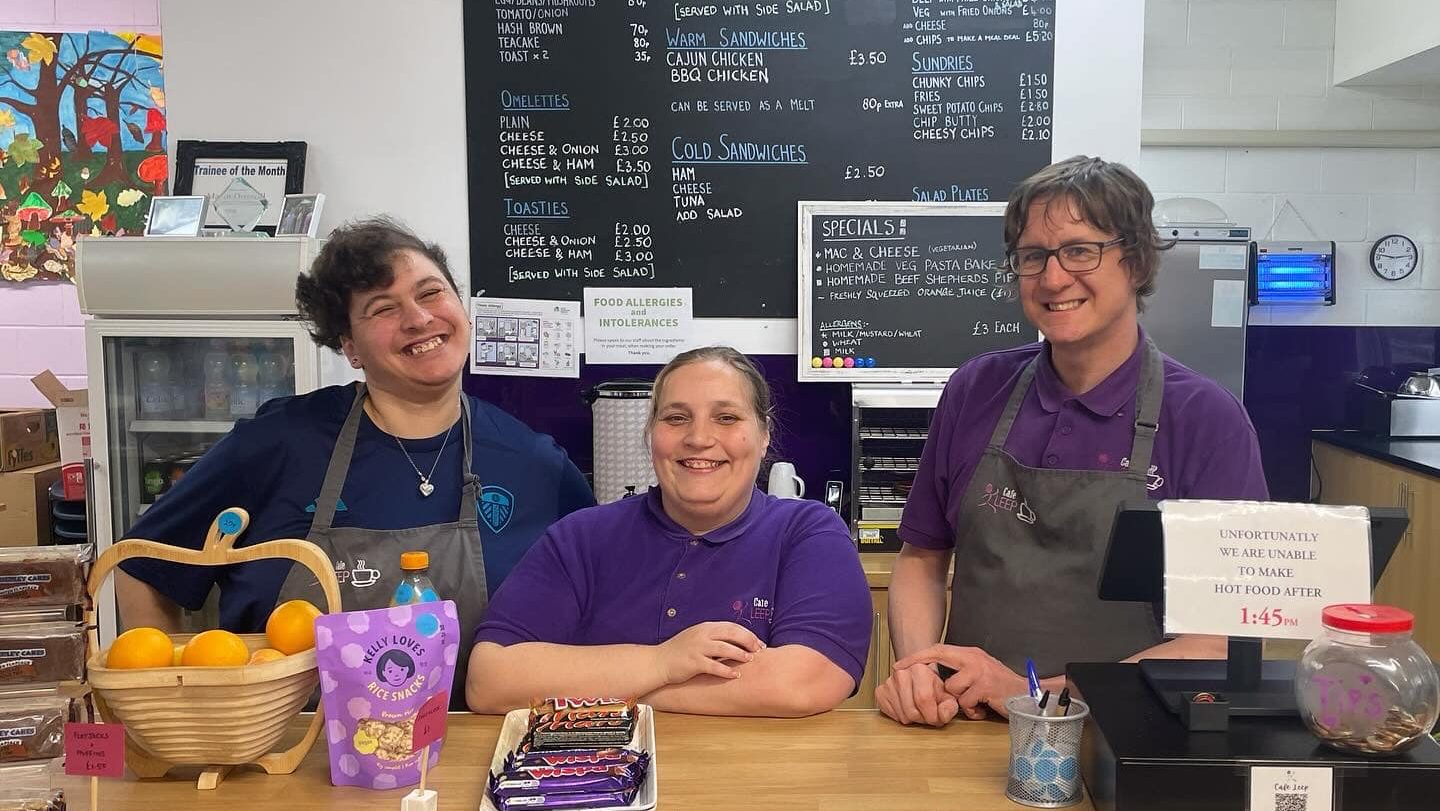A photo of staff and trainees from Cafe Leep, stood behind a counter and in front of a chalkboard with their menu. They are all wearing purple polo shirts and two of them are wearing dark grey aprons. They are all smiling.