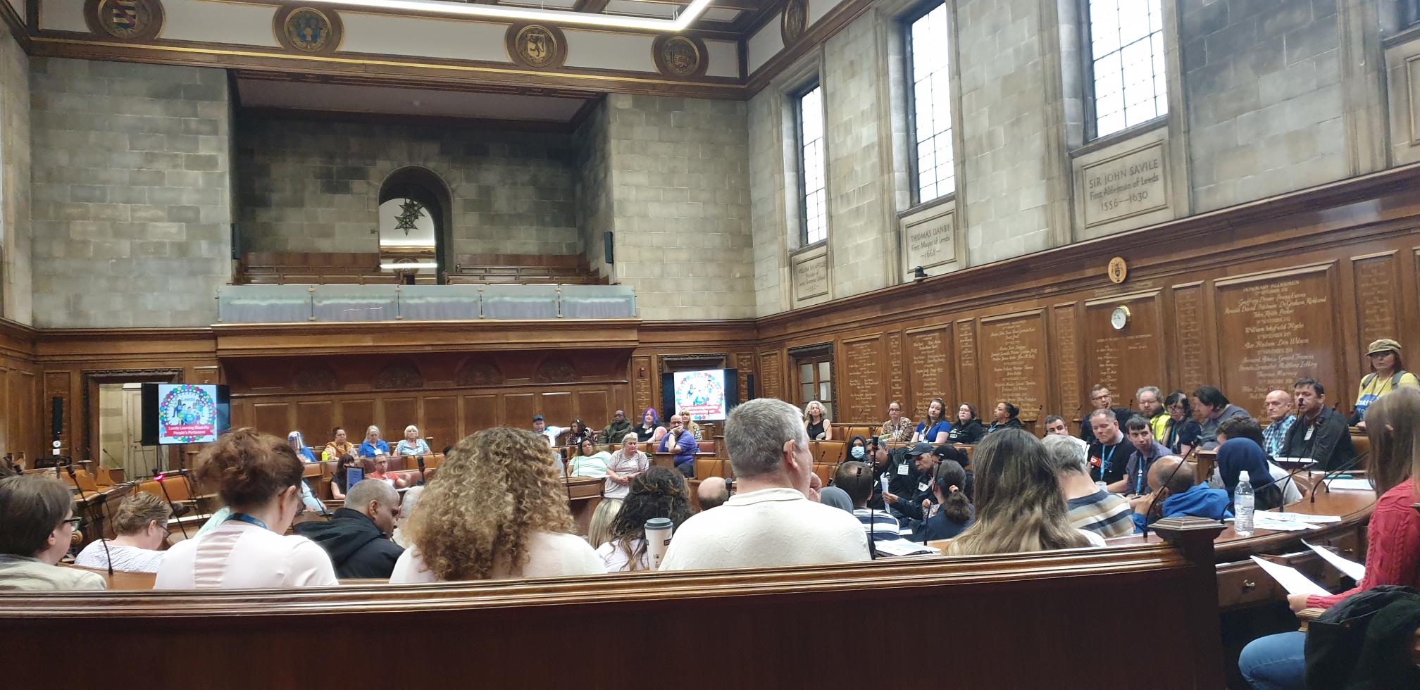 An image from the 2023 Council Chamber Takeover. There are people sat in a room in Leeds Civic Hall, in circular rows of seats facing a screen.