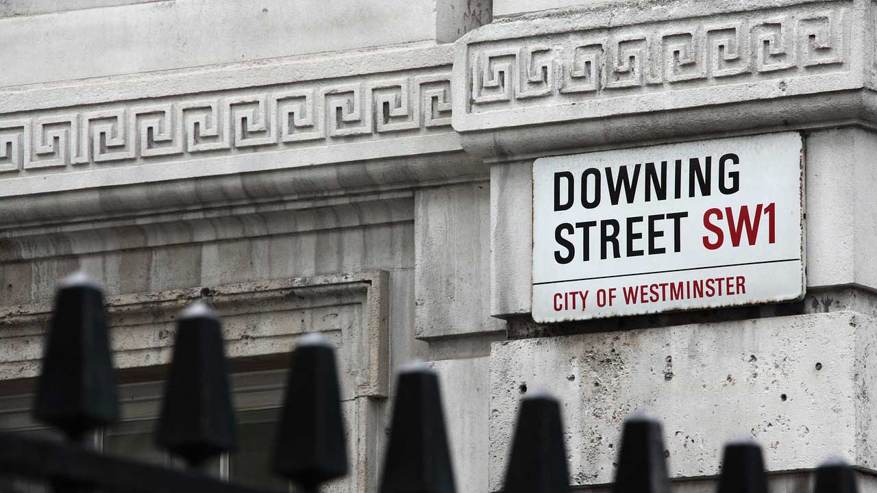 An image of the street sign for Downing Street. The sign is on a stone wall, with the top of some black metal railings in front of it.