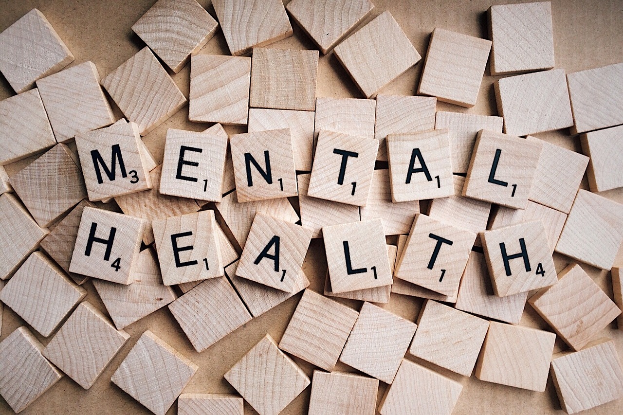 The words 'Mental Health' spelled out using wooden tiles with each letter on one tile. They are on a bed of blank wooden tiles.