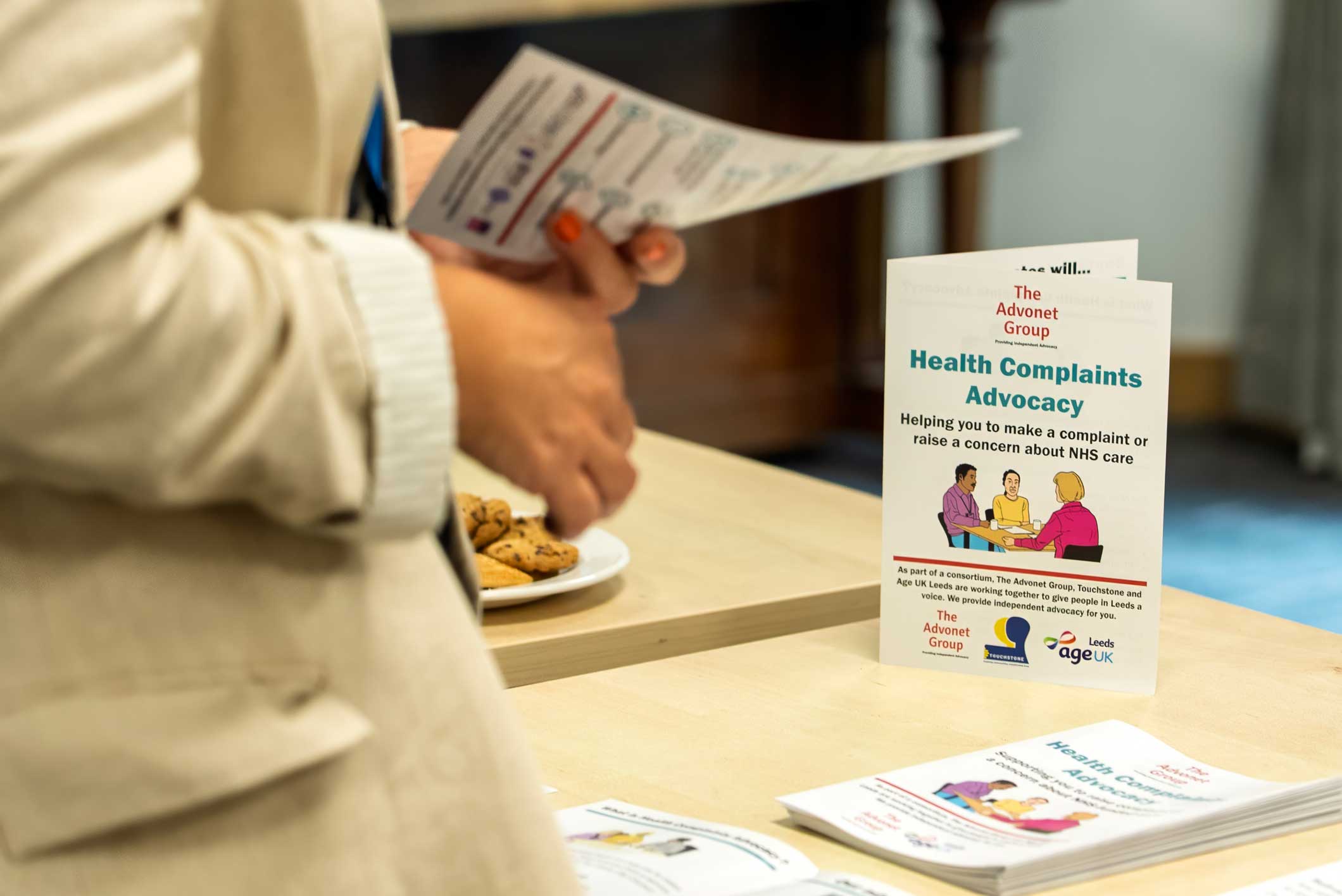 A photo of someone holding a leaflet in their hand. In the background, there is a leaflet for Health Complaints Advocacy stood on a table.