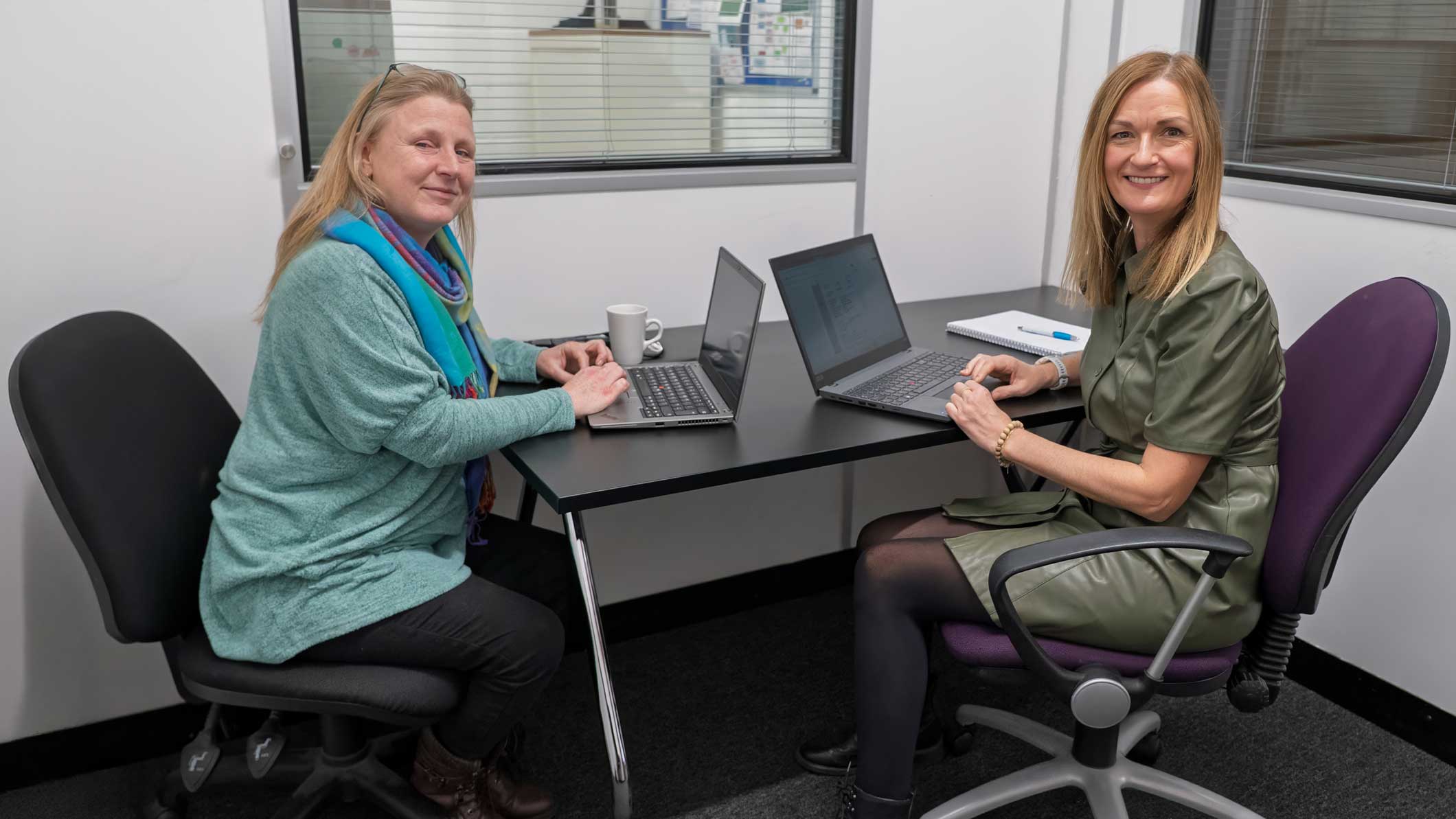 A photo of two members of staff sat at a desk, facing the camera and smiling