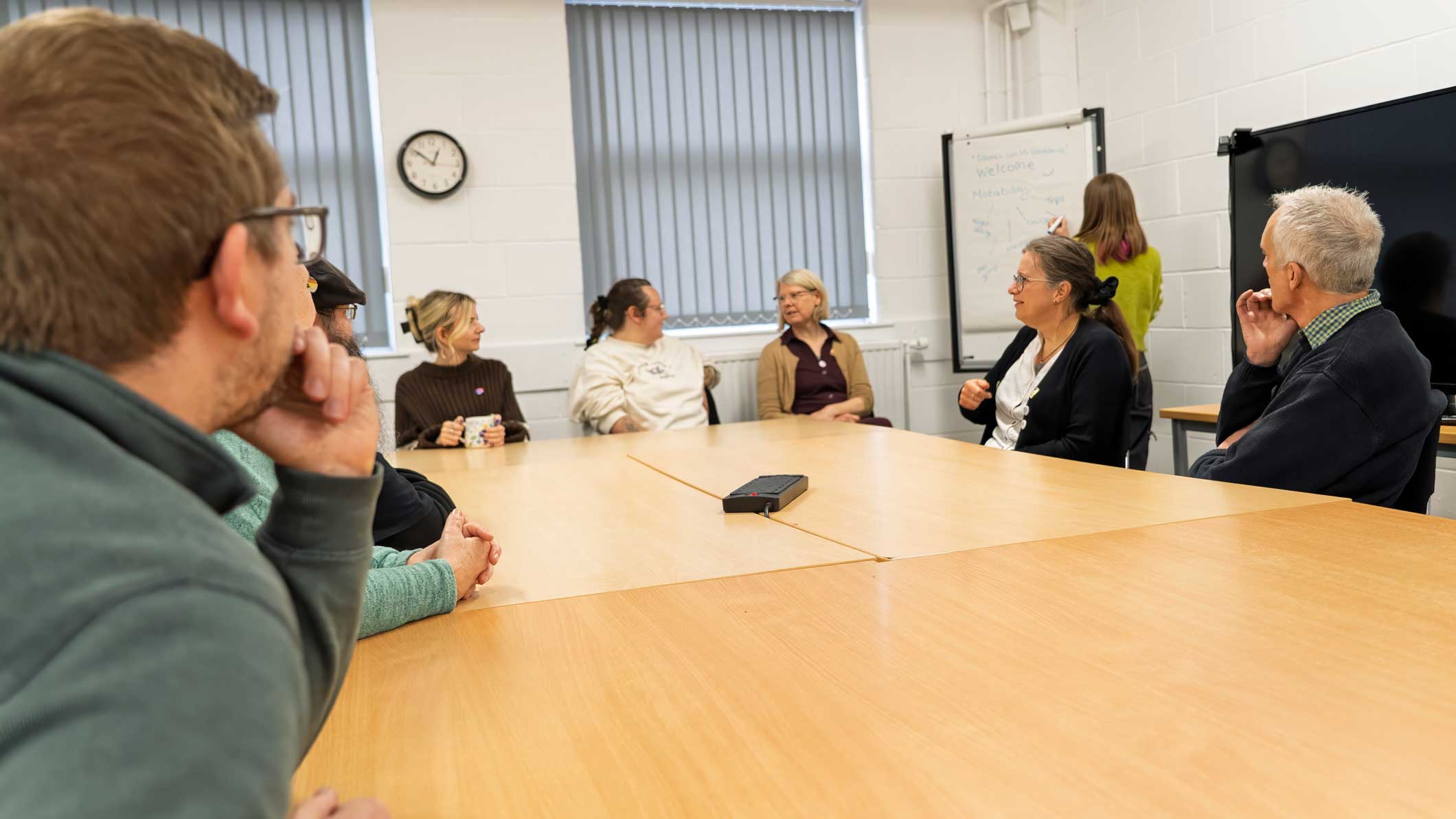 A photo of members of our staff team in a meeting, sat around a wooden table. One staff member is stood next to a flipchart, writing on it.