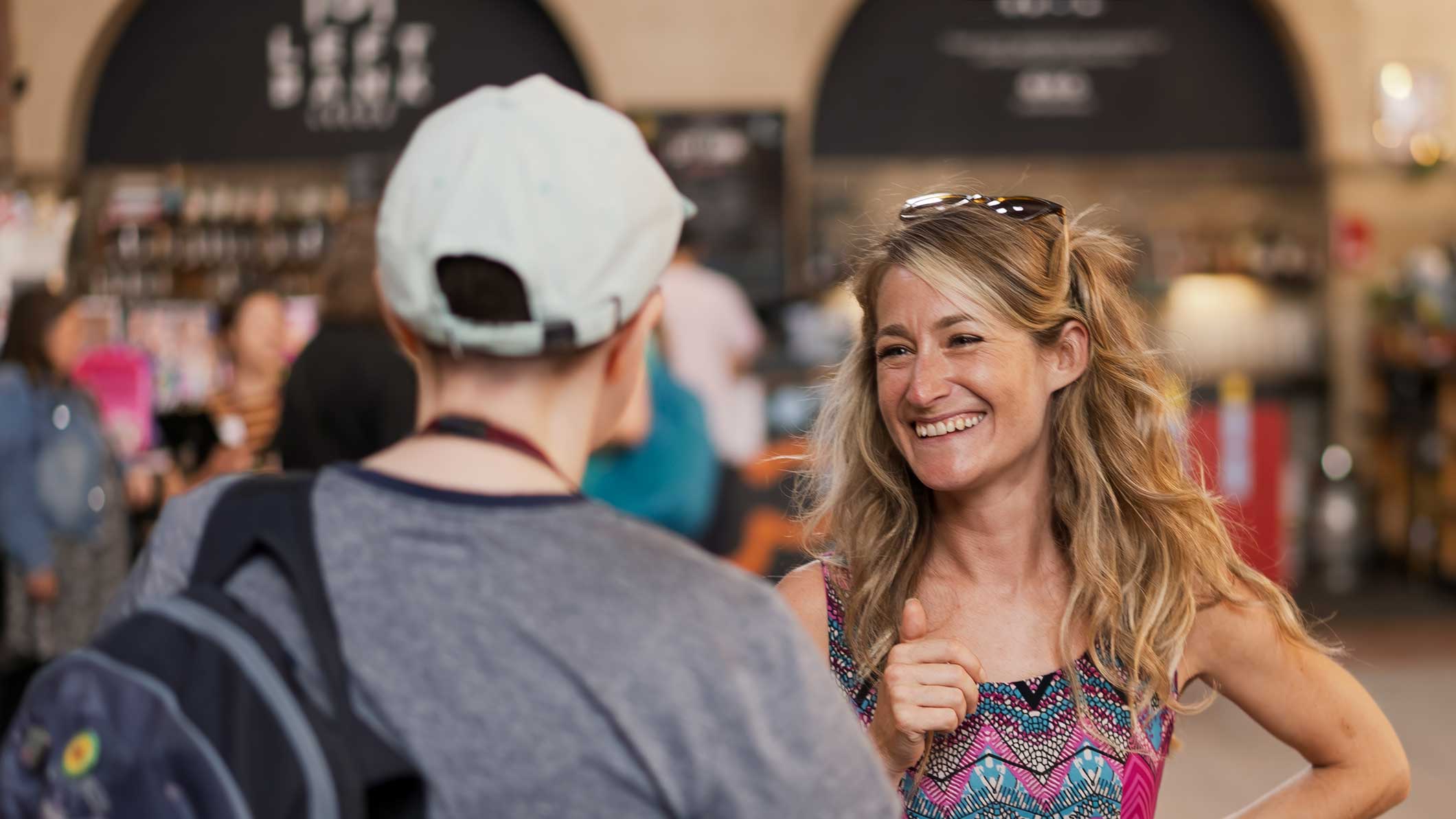 A photo of two members of our staff team talking to each other at an event. One of them has their back facing the camera and is wearing a grey t-shirt and cream baseball cap. The other has blonde hair and is wearing a blue, pink, black and white patterned dress.